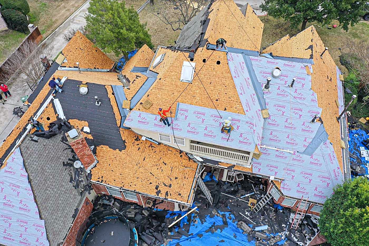 Drone view of a large brick home part way through a tear-off, with decking exposed and new underlayment down on one plane