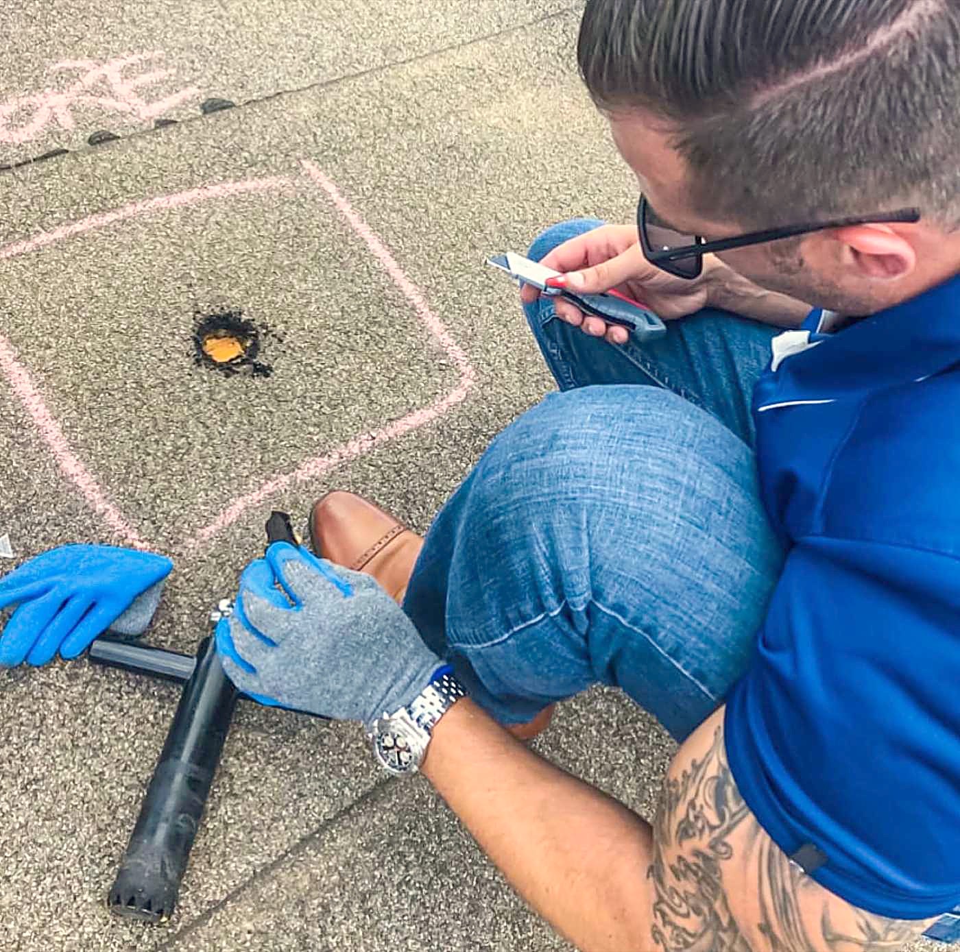 A hand marking a chalk test square around a hail strike on an asphalt shingle roof, with a measuring tool alongside